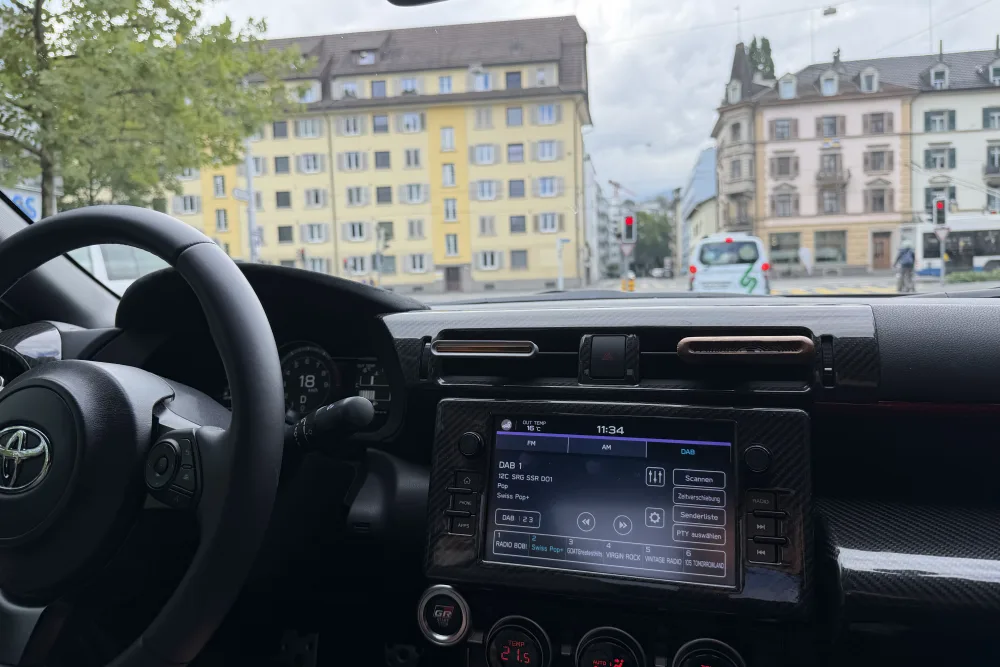Blick vom Fahrersitz durch die Windschutzscheibe auf eine Kreuzung mit roter Ampel; im Vordergrund Lenkrad, Armaturenbrett und Radio.