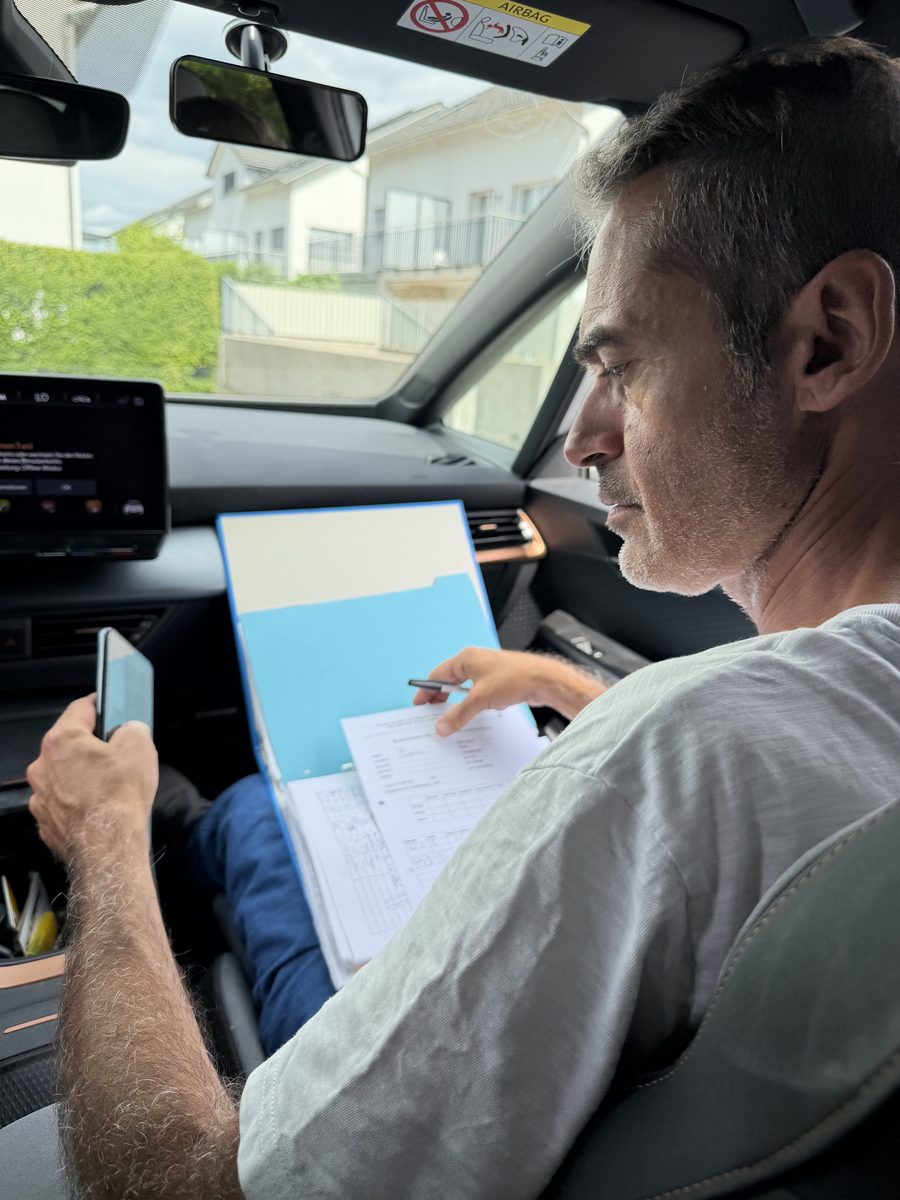 Ein Fahrlehrer auf dem Beifahrersitz füllt nach der Fahrstunde einen gedruckten Bewertungsbogen auf einer blauen Mappe aus.