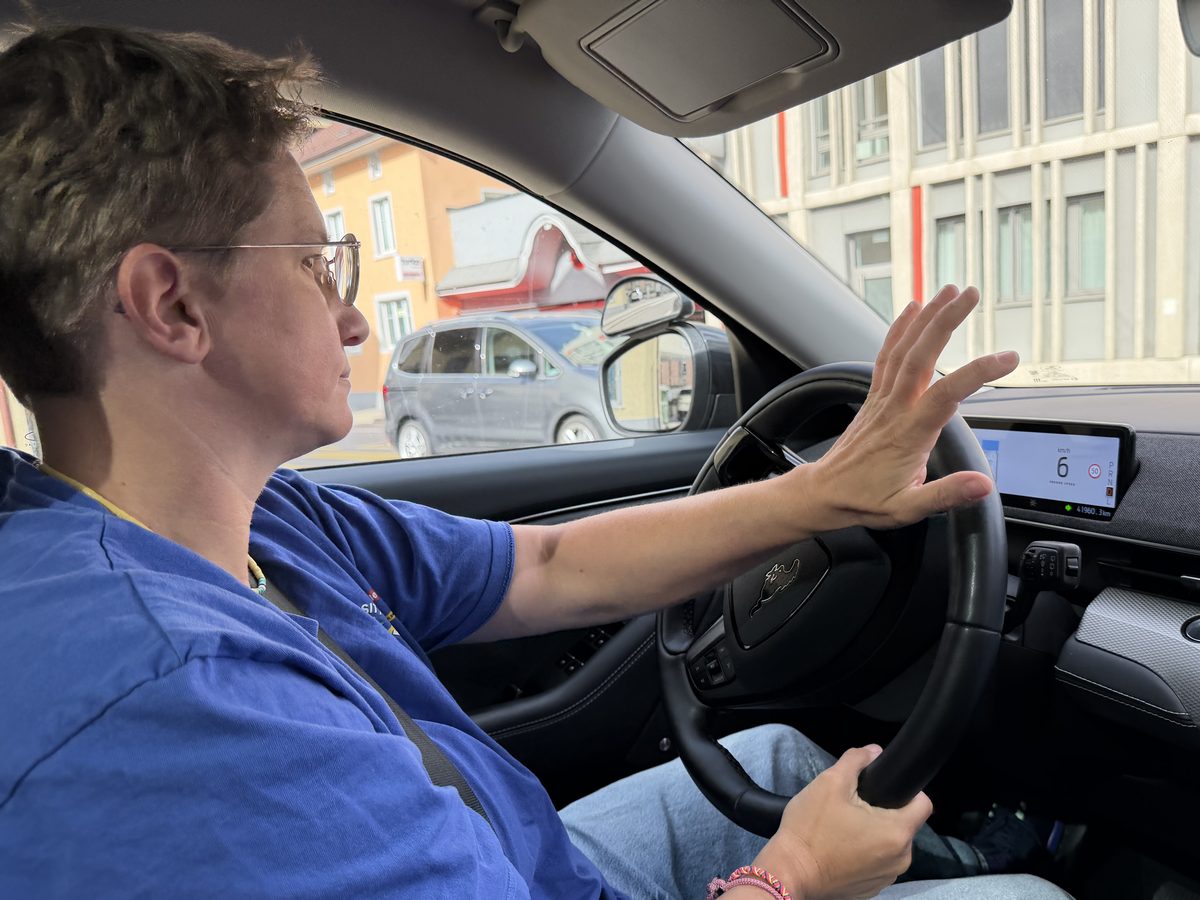 Eine Person mit Brille und blauem T-Shirt am Steuer eines Autos, eine Hand am Lenkrad, die andere erhoben. Durch die Seitenscheibe ein oranges Haus und ein graues Auto.
