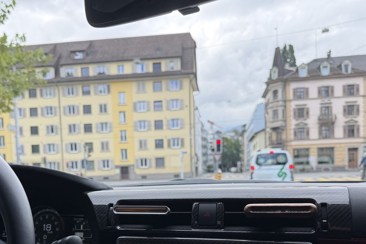 Blick durch die Windschutzscheibe auf eine Strasse in einer Stadt: links ein gelbes, rechts ein rosa Haus, in der Mitte eine rote Ampel, vorne ein weisses Auto, unten das Armaturenbrett.