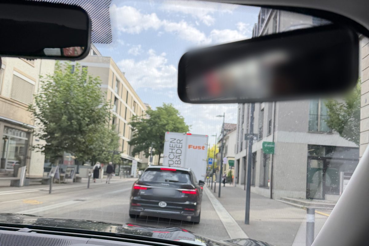 Blick durch die Windschutzscheibe auf eine Strasse in einer Stadt: vorne ein dunkles Auto und ein weisser Lastwagen, rechts ein Trottoir, auf beiden Seiten Häuser und Bäume.